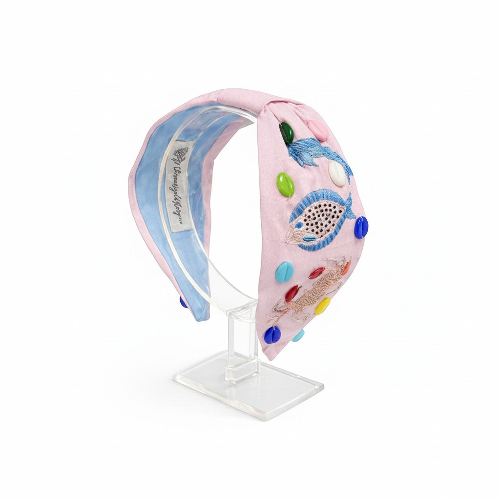 The Marigold Story pink headband with hand-embroidered fish, seashell accents, and colorful beads on a blush base.