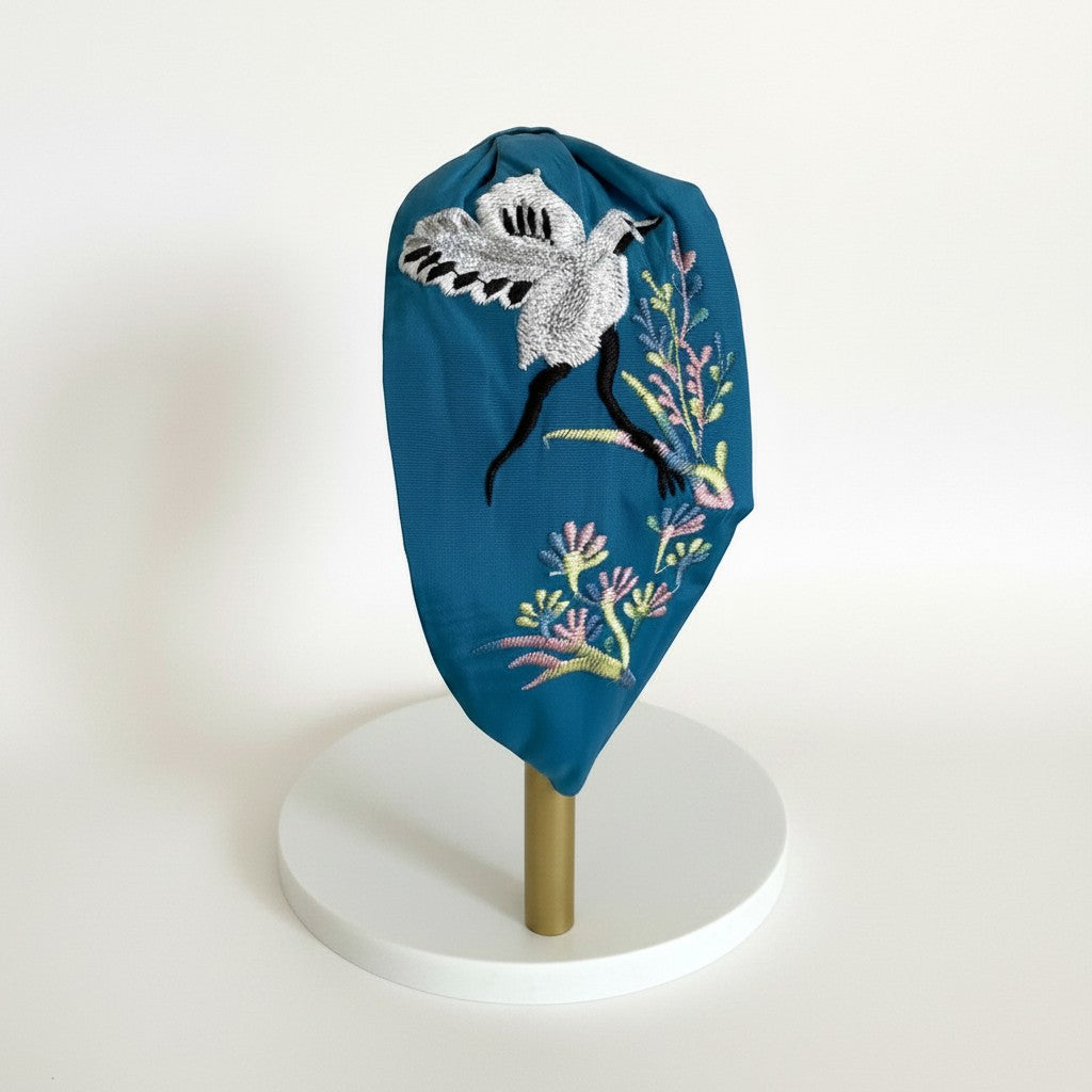 The Marigold Story teal padded headband featuring hand-embroidered silver crane and pastel botanical floral motifs.