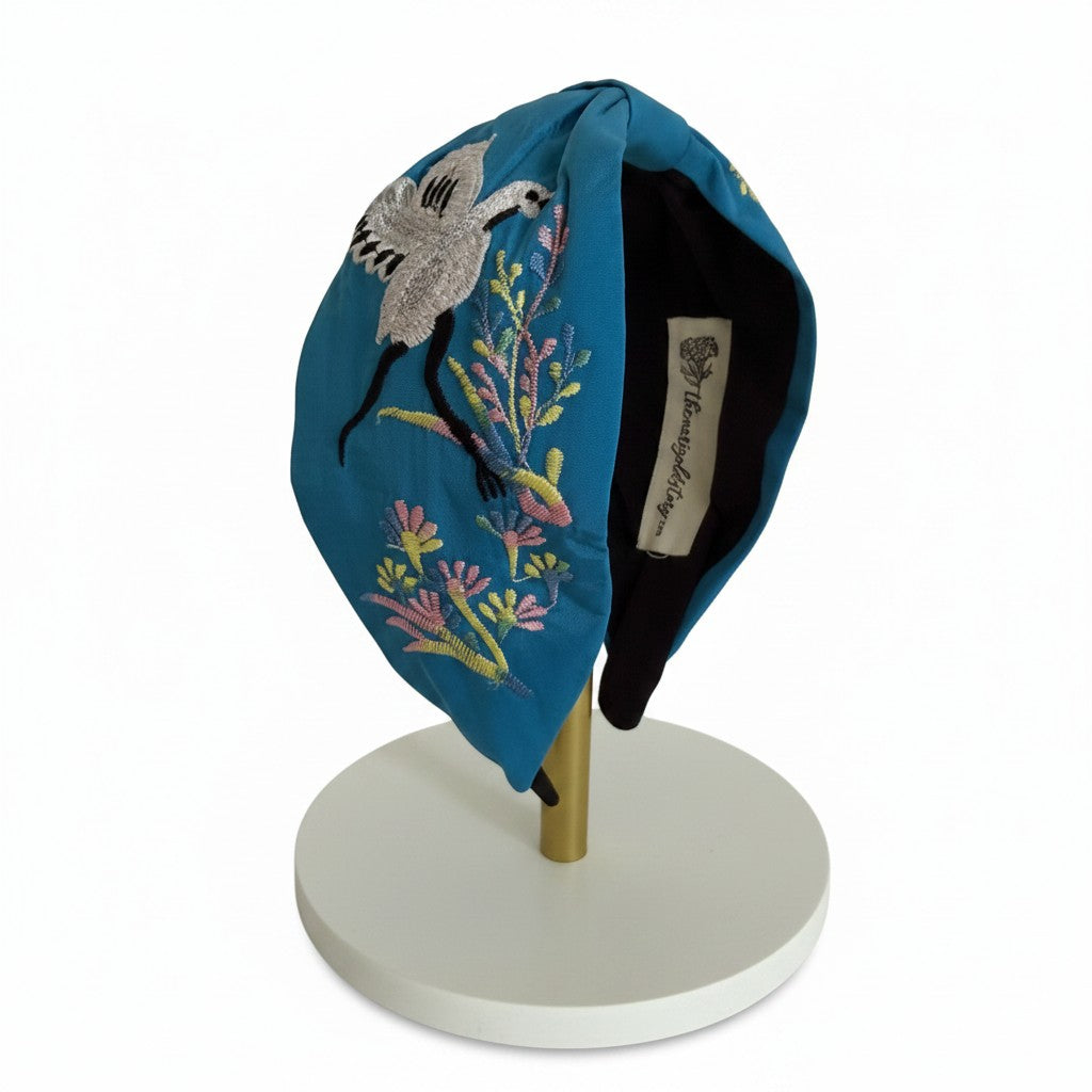 The Marigold Story teal padded headband featuring hand-embroidered silver crane and pastel botanical motifs.